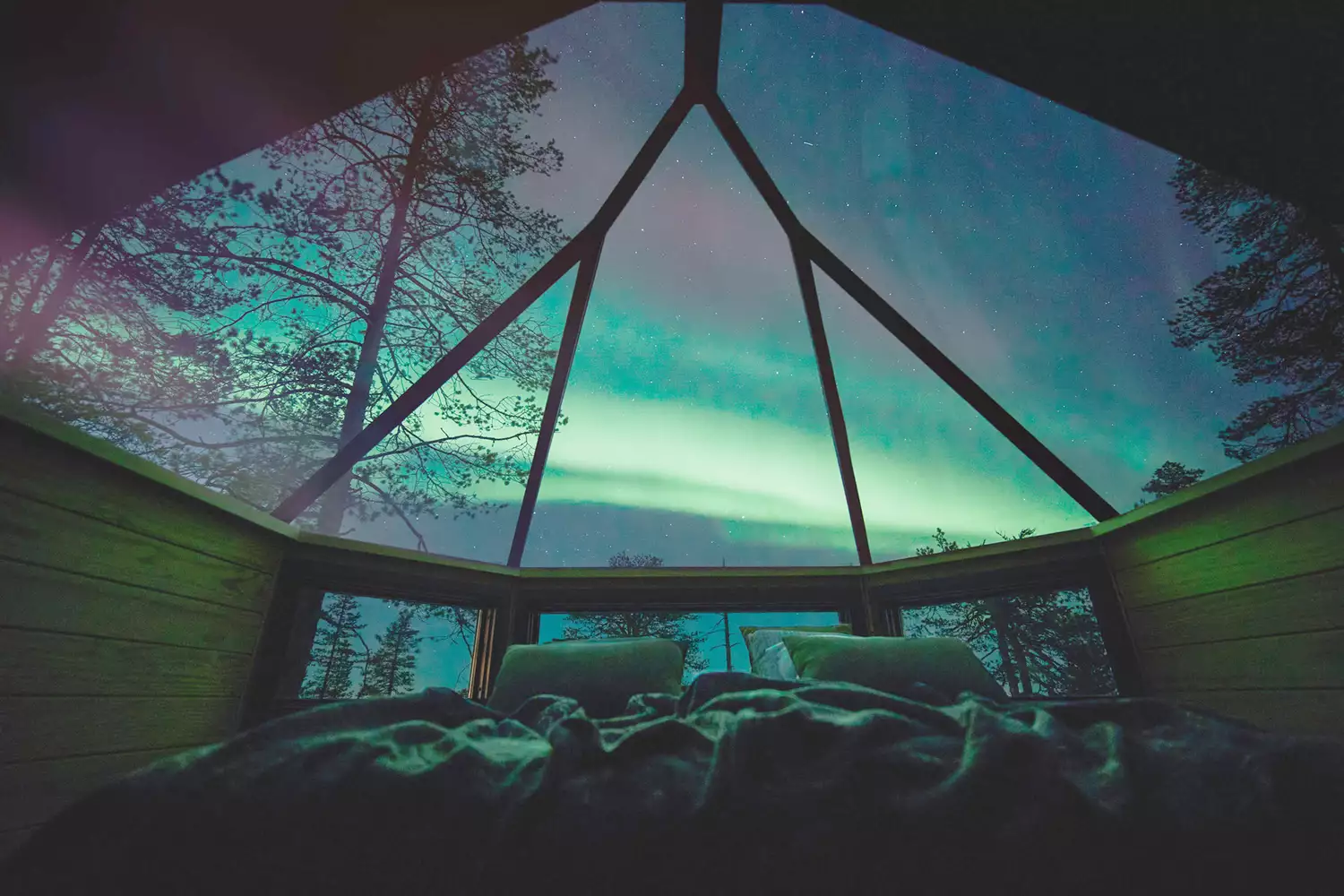 A bed looking through a glass ceiling of an igloo at northern lights